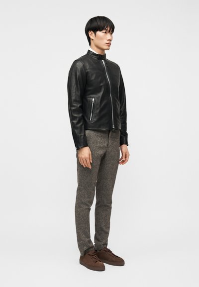 Άνδρας όρθιος, φοράει μαύρο δερμάτινο μπουφάν με φερμουάρ, γκρι υφασμάτινο παντελόνι και καφέ αθλητικά παπούτσια με κορδόνια, σε απλό φωτεινό φόντο.