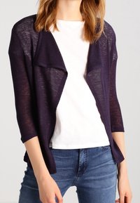 Cardigan leggero, aperto sul davanti, di colore viola scuro, con maniche a tre quarti e motivo a maglia strutturata, indossato sopra una camicia bianca e jeans blu.