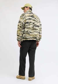 Fuzzy bež i crni prugasti kaput, uparen s tamnim hlačama i smeškim čizmama, uz laganu zelenu kapu. Tekstura, opušteni kroj.