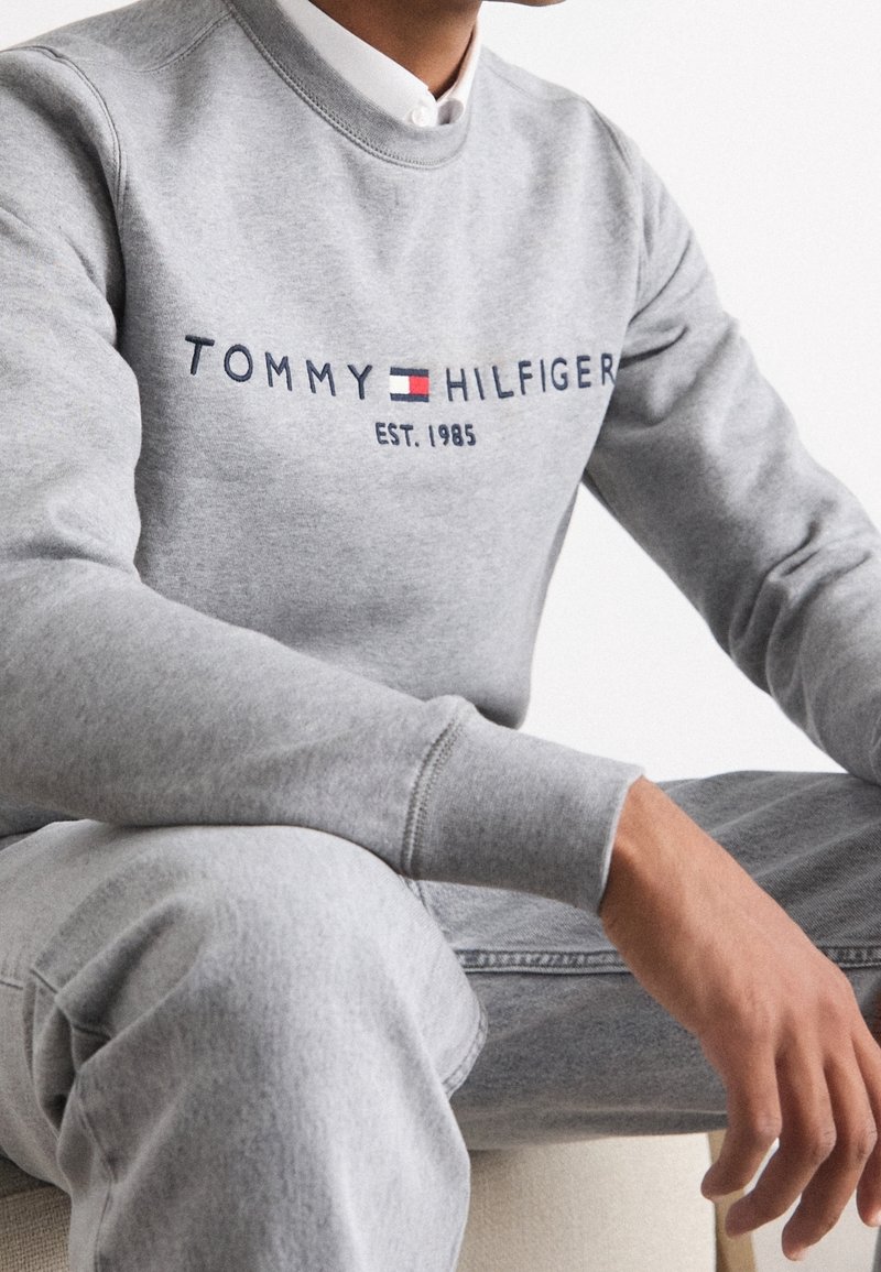 Personne portant un sweat-shirt Tommy Hilfiger gris clair et un jean, assise avec les mains posées sur les genoux, texte visible "EST. 1985" sur le sweat-shirt.