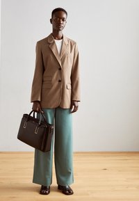 Lysebrun skræddersyet blazer, grønne bukser med vide ben, sort håndtaske med gulddetaljer og sorte sandaler med blokhæl. Glat tekstur hele vejen igennem.