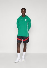 Grøn hoodie med lynlås og logo, sorte shorts med rød stribe, hvide sko og sokker. Modellen står mod en ensfarvet baggrund.