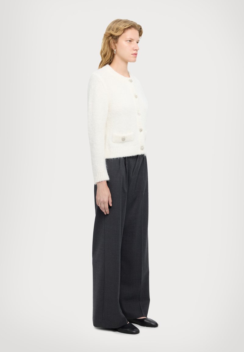 Weißer flauschiger Cardigan mit Perlenknöpfen, zwei Vorderaschen, kombiniert mit dunkelgrauen weit geschnittenen Hosen und schwarzen Slippern. Seitenansicht.