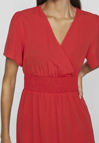 Červené šaty s krátkým rukávem ve stylu wrap z lehkého materiálu. Mají smockovaný pas a výstřih do V, s volnou sukní a měkkou strukturou.