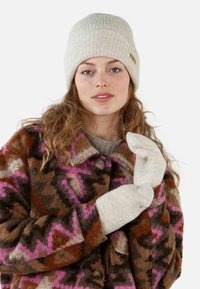 Mujer con un sombrero de punto beige, manoplas a juego y un abrigo de peluche de colores marrón, rosa y negro con un patrón, ajustando una de las manoplas.