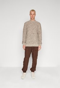 Šviesiai rudos spalvos megztinis su tekstūruotu rombo raštu, derinamas su rudais krovinių tipo kelnėmis, turinčiomis kelias kišenes, ir béžiniais sportbačiais.