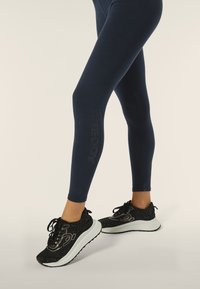 Leggings blu navy con logo in rilievo sul lato, abbinati a sneaker nere con tomaia maculata e suola bianca.