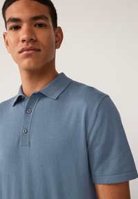 Polo shirt blu con una trama morbida, caratterizzato da un colletto classico, maniche corte e una patta con tre bottoni. Primo piano dell'indumento superiore.