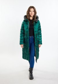 Cappotto imbottito lungo e trapuntato di colore verde scuro con cappuccio, dotato di zip frontale e due tasche laterali. Indossato sopra un top nero e jeans blu.