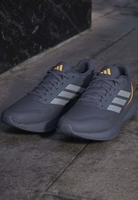 Chaussures de course Adidas gris foncé avec trois bandes réfléchissantes et accents de logo orange, placées côte à côte sur un pavé en pierre.