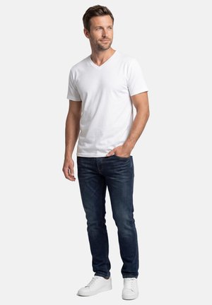 Man standing with one hand in pocket, wearing a white V-neck t-shirt, dark jeans, and white sneakers, looking slightly to the side.