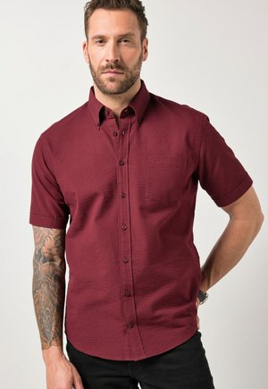 Mężczyzna z krótką brodą w teksturowanej, zapinanej na guziki bordowej koszuli z krótkim rękawem, pokazujący szczegółowy tatuaż na lewej ręce.