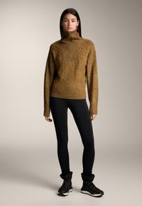 Un pull en tricot marron avec un col haut, un motif texturé et une coupe ajustée, associé à des leggings noirs et des bottes à lacets noires.