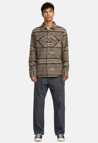 Camicia con bottoni caratterizzata da un motivo a righe marroni e blu, due taschini sul petto e una vestibilità leggermente rilassata, abbinata a pantaloni grigio scuro e sneakers.