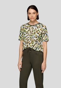 Frau mit kurzem, dunklem Haar, die ein kurzärmliges, mehrfarbiges Leopardenmuster-Oberteil und dunkle grüne Hosen mit Reißverschlusstaschen trägt.