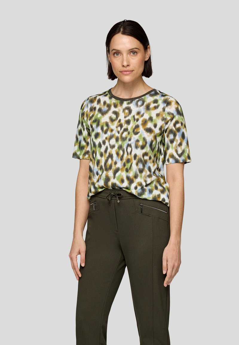 Frau mit kurzem, dunklem Haar, die ein kurzärmliges, mehrfarbiges Leopardenmuster-Oberteil und dunkle grüne Hosen mit Reißverschlusstaschen trägt.