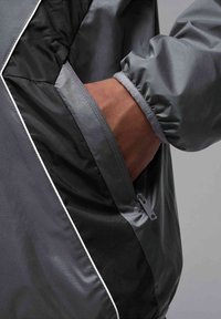 Grå vindjacka med elastiska ärmmuddar, kontrasterande svart foder, ficka med dragkedja och reflekterande vit kant längs sömmarna.