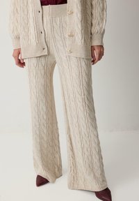 Lichtbeige gebreide trui en wijde broek met een gestructureerd kabelpatroon. De outfit is gecombineerd met donkerrode puntige schoenen.