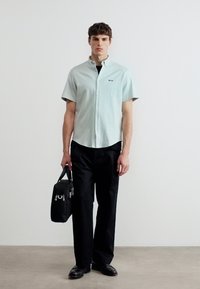 Νέος άνδρας που φοράει ανοιχτόχρωμο πουκάμισο με κοντό μανίκι, μαύρο παντελόνι και παπούτσια, κρατώντας μαύρη τσάντα προσώπου πάνω σε απλό λευκό φόντο.