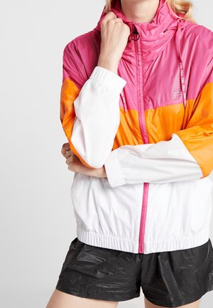 Person wearing a pink, orange, and white color-block windbreaker jacket with black shorts, arms crossed and one hand near the face.