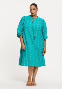 Türkises Baumwollkleid mit Puffärmeln, einem Schlüssellochausschnitt und Quasten-Detail. Verfügt über florale Muster. Die Länge reicht über das Knie.