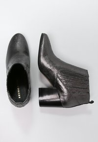 Schwarze Knöchelschuhe mit einer strukturierten, reptilartigen Oberfläche. Mit blockartigem Absatz und Fransenverzierungen an der Seite. Spitz zulaufendes Design.