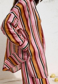 Camicia a righe leggera nei toni del rosa, arancione e nero. Presenta una vestibilità ampia, colletto con bottoni e maniche lunghe con polsini a contrasto.