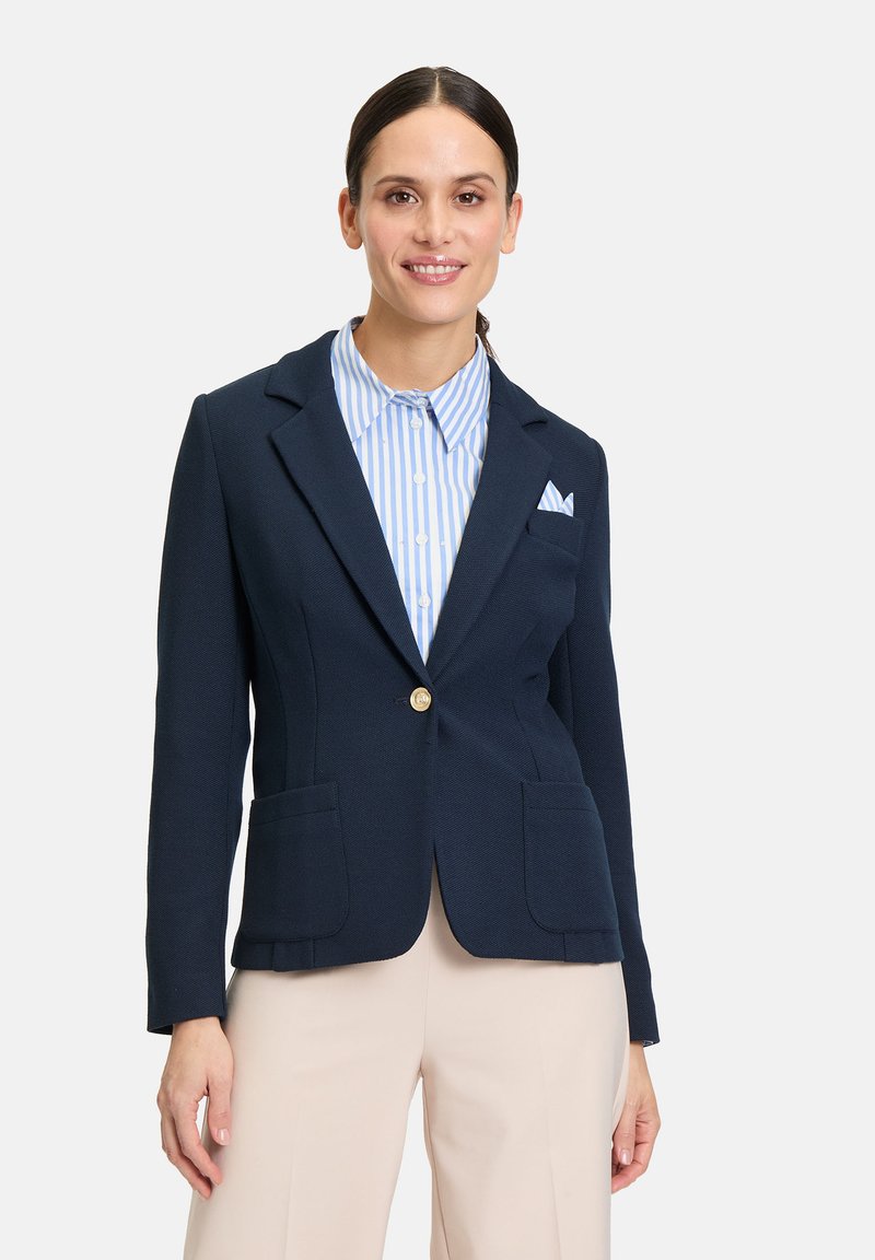 Granatowa marynarka o fakturowanym wykończeniu, z jednym złotym guzikiem, dwiema przednimi kieszeniami oraz białą koszulą w niebieskie paski pod spodem.