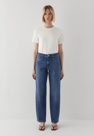 Blonde Frau steht, trägt ein weißes T-Shirt mit kurzen Ärmeln, hoch taillierte gerade blaue Jeans und braune Sandalen vor einem einfarbigen hellen Hintergrund.