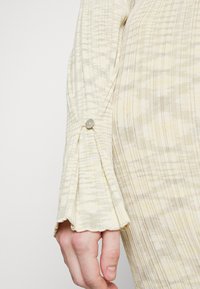 Gros plan d'une main portant une manche longue en côtes beige avec un poignet boutonné et un motif à carreaux subtil.