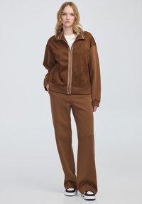Giacca marrone con zip, dal texture scamosciato e polsini a costine, abbinata a pantaloni della tuta a gamba larga e scarpe con suola bianca.