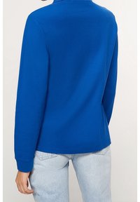 Niebieska bluzka z długim rękawem, wykonana z miękkiego materiału, o prostym fasonie, okrągłym dekolcie i mankietach, zestawiona z jasnoniebieskimi dżinsami.