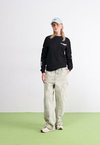 Camisa negra de manga larga con un gráfico en la parte frontal, combinada con pantalones holgados de color verde claro y una gorra azul clara. Tela suave, ajuste casual.