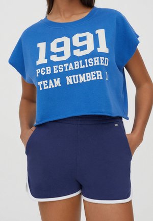 Kobieta ubrana w niebieski krótki top bez rękawów z napisem „1991 P&B Established Team Number” oraz granatowe sportowe spodenki z białym obszyciem.