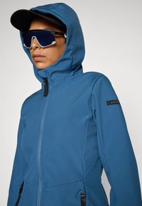 Blå softshell-jakke med glidelås foran, justerbar hette og logo på ermet. Har en jevn tekstur og minimale hardware-detaljer.
