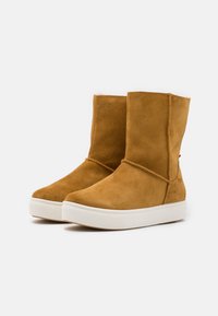 Bottines en daim marron clair avec une semelle blanche texturée. Présentent un design unique avec des coutures croisées et une doublure intérieure douce.