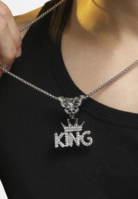 Sølv kædedesign med en sommerfugl som forbindelsesled og en rhinsten "KING" vedhæng med en kroneaccent, båret over en sort top.