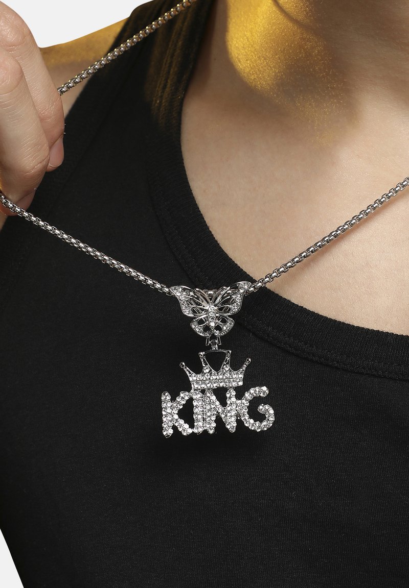 Sølv kædedesign med en sommerfugl som forbindelsesled og en rhinsten "KING" vedhæng med en kroneaccent, båret over en sort top.