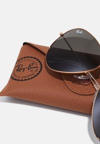 Ochelari de soare Ray-Ban într-un capac texturat maro. Prezintă lentile închise la culoare, ramă din metal aurie și logo-ul imprimat în negru pe capac.