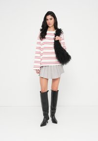 Μπλούζα με μακριά μανίκια κόκκινη με λευκές ρίγες, γκρι πλισέ φούστα, μαύρες μπότες μέχρι το γόνατο και μια αφράτη μαύρη τσάντα. Απλό φόντο.