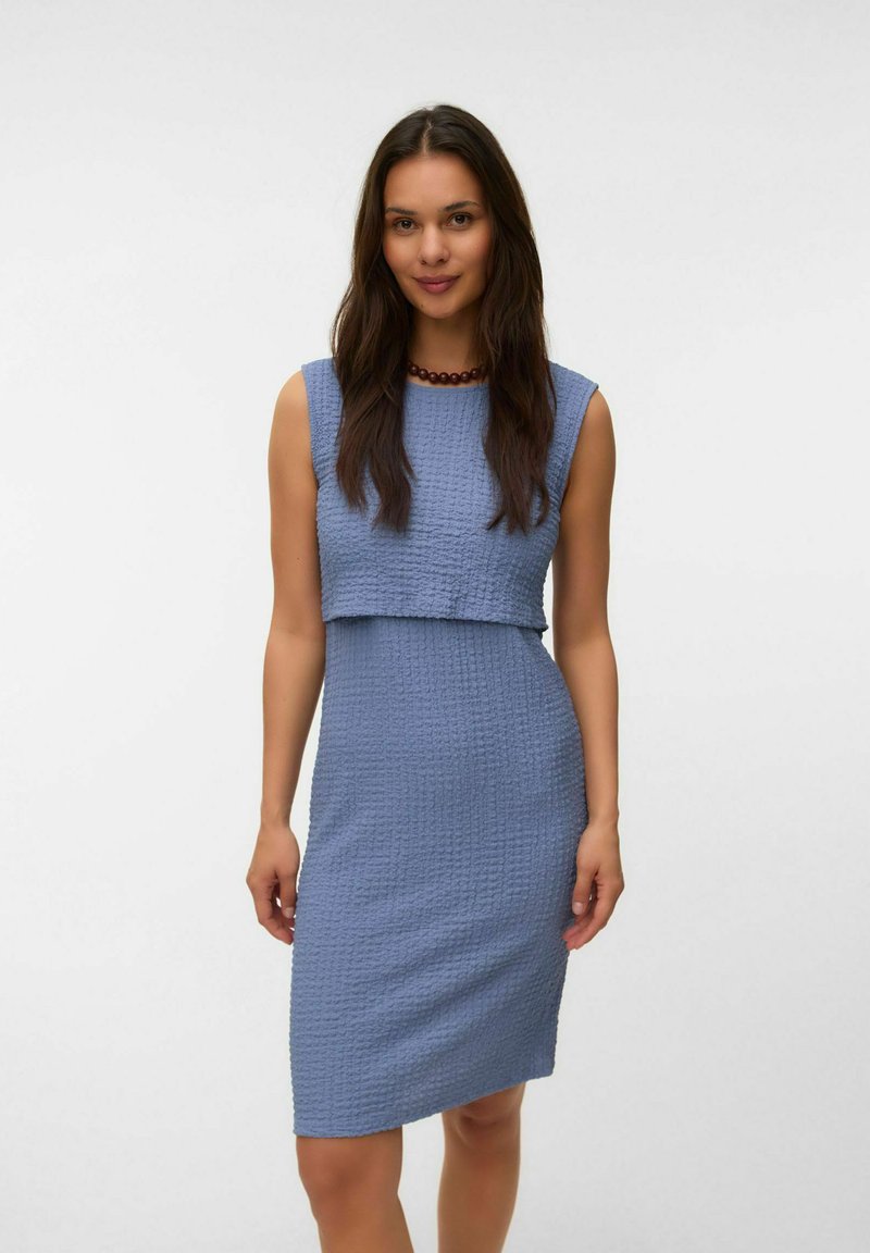 Femme aux longs cheveux portant une robe bleue texturée sans manches et un collier sombre à perles, debout devant un fond blanc uni.