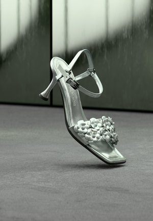 Silberne High-Heel-Sandale mit Knöchelriemen und Blumendekorationen am vorderen Riemen, vor neutralem Hintergrund gezeigt.