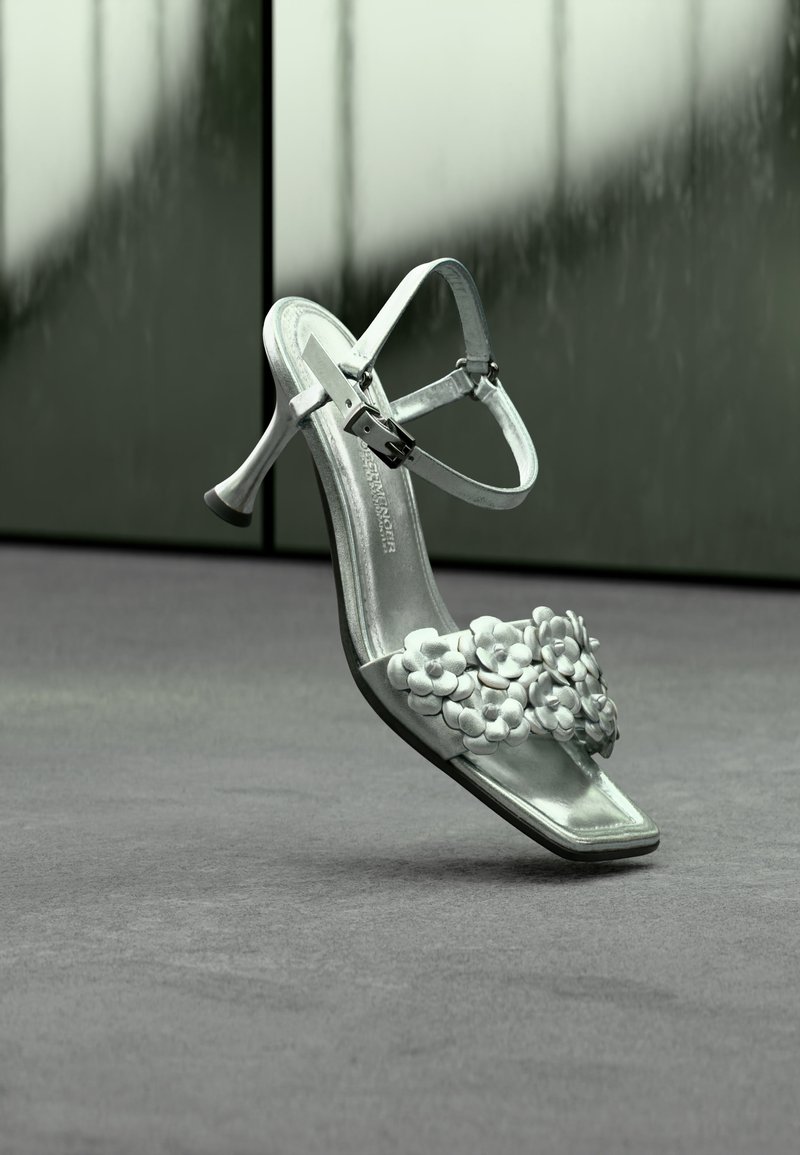 Silberne High-Heel-Sandale mit Knöchelriemen und Blumendekorationen am vorderen Riemen, vor neutralem Hintergrund gezeigt.