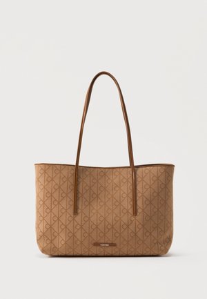 Smeđa tote torba Calvin Klein s monogram uzorkom i tankim naramenicama na jednobojnoj svijetloj pozadini.