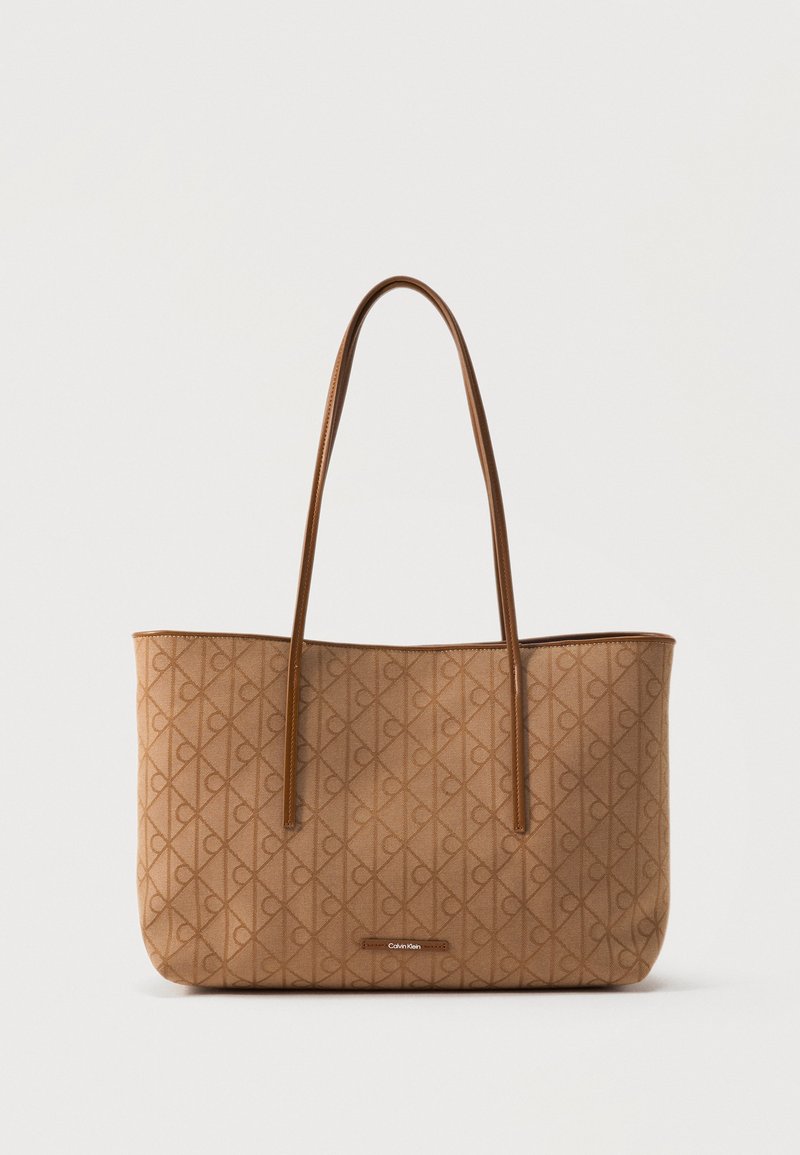 Καφέ τσάντα tote Calvin Klein με μοτίβο μονόγραμμα και λεπτούς ιμάντες ώμου σε απλό φωτεινό φόντο.