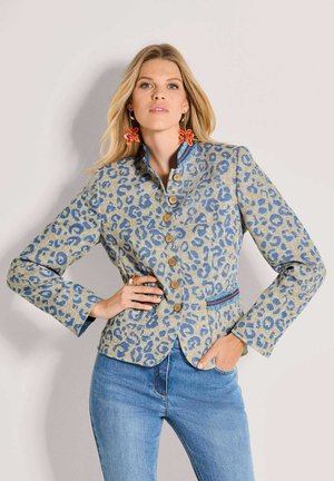 Frau trägt einen blau gepunkteten Blazer mit Leopardenmuster, blaue Jeans, orange Blumenohrringe, eine Hand in der Hüfte und die andere in der Tasche.