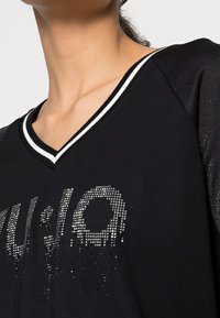 Zbliżenie osoby noszącej czarną bluzkę z dekoltem w serek, ozdobioną białą lamówką w paski i srebrnymi literami z otoczkami z cyrkonii na piersi.