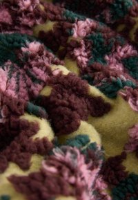 Textil s textúrou zobrazujúci zoskupenia kvetinových vzorov v ružovej, tmavozelenej a tmavobordovej farbe na žltom pozadí. Mäkký a nadýchaný vzhľad.