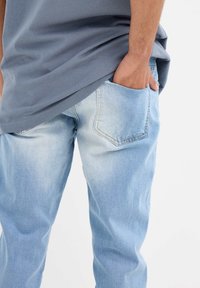 Jean de mezclilla azul claro con una textura ligeramente desteñida, que cuenta con un bolsillo trasero y un corte relajado, combinado con una camiseta gris.
