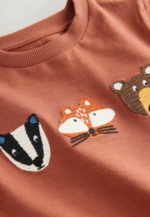 Roodbruine t-shirt met geborduurde dierengezichten: een zwart-witte das, een vos met oranje accenten en een bruine beer. Zachte stof.